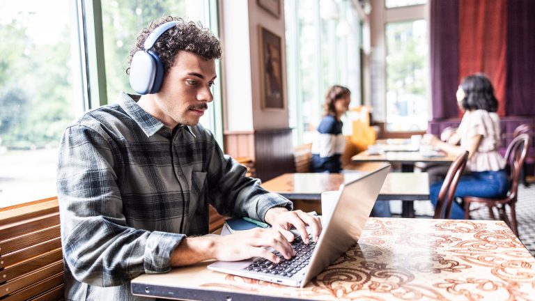 Eine Person sitzt mit Apple AirPods Max auf dem Kopf an einem MacBook Pro in einem Caf&eacute;.