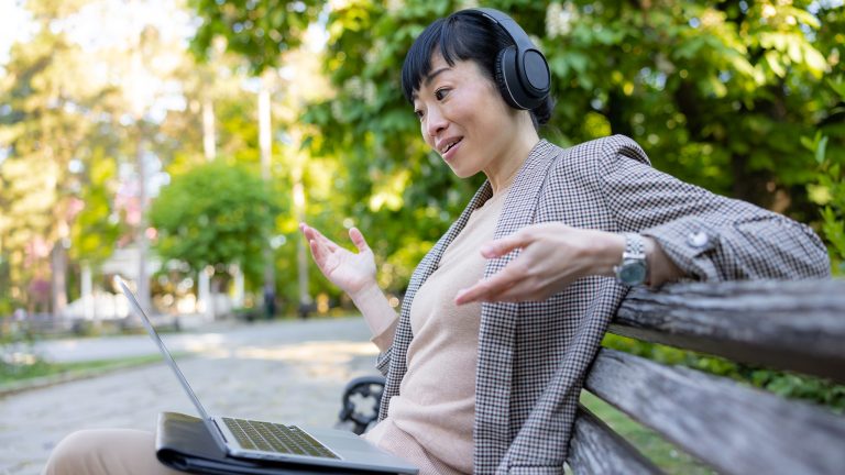 Eine Person mit Over-Ear-Kopfh&ouml;rern sitzt auf einer Bank und scheint &uuml;ber ihren Laptop ein Meeting zu halten.