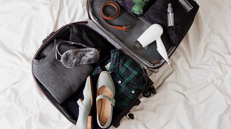 (Ein Bild von einem Reisef&ouml;hn, der zusammen mit Schuhen, einem G&uuml;rtel, einer Schlafmaske und mehr in einem ge&ouml;ffneten Koffer auf einem Bett liegt.)
