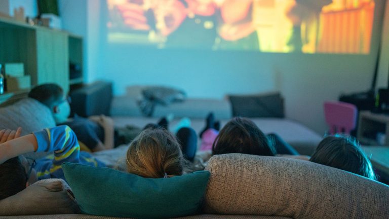 F&uuml;nf Personen liegen auf einem Sofa und schauen in Richtung eines Beamer-Bildes an die Wand.