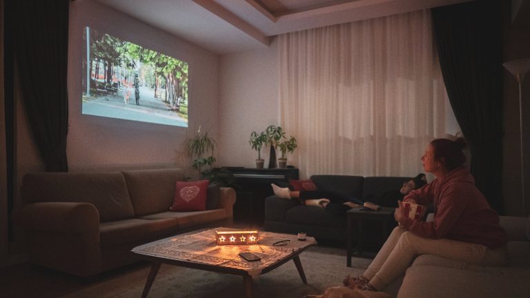 Zwei Personen sitzen und liegen auf einer Wohnlandschaft in einem abgedunkelten Wohnzimmer und schauen einen Film, der von einem Beamer auf eine Wand projiziert wird.