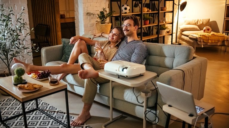 Zwei Personen sitzen Arm in Arm auf einem Sofa in einem gem&uuml;tlich eingerichteten Wohnzimmer. Auf einem kleinen Beistelltisch steht ein Beamer.