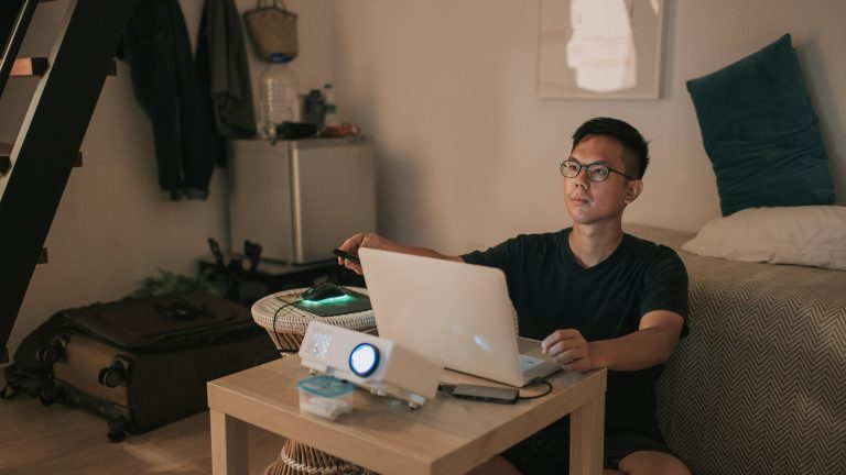 Eine Person sitzt im Schneidersitz vor einem Wohnzimmertischchen auf dem ein Laptop steht, der an einen Beamer angeschlossen ist.