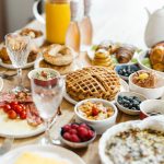 Auf einem Tisch stehen viele verschiedene Speisen f&uuml;r einen Brunch, wie Waffeln, Heidelbeeren, Bagels, Spiegeleier und Sektgl&auml;schen.