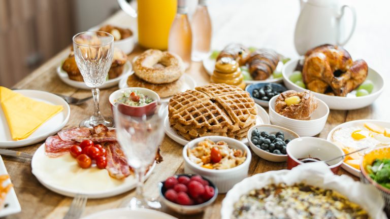 Auf einem Tisch stehen viele verschiedene Speisen f&uuml;r einen Brunch, wie Waffeln, Heidelbeeren, Bagels, Spiegeleier und Sektgl&auml;schen.
