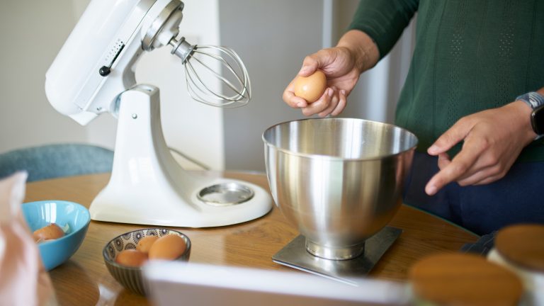 Es sind die H&auml;nde einer Person zu sehen, die ein Ei am Rand der Sch&uuml;ssel einer K&uuml;chenmaschine, welche im Hintergrund steht, zerschl&auml;gt.