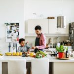 Eine Person und ein Kind stehen in einer offenen, hellen K&uuml;che. In der Mitte steht eine K&uuml;cheninsel, auf der sie gerade Obst und Gem&uuml;se schneiden.