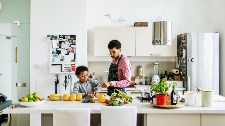 Eine Person und ein Kind stehen in einer offenen, hellen K&uuml;che. In der Mitte steht eine K&uuml;cheninsel, auf der sie gerade Obst und Gem&uuml;se schneiden.