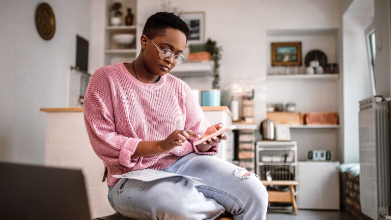 Eine Person sitzt in einer Wohnung auf einem Tisch und nutzt ein Smartphone. Im Hintergrund sind Regale und eine offene K&uuml;che zu sehen.
