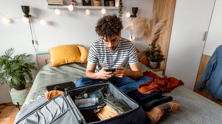 Eine Person sitzt auf einem Bett und schaut auf ein Smartphone. Vor der Person liegt ein offener Koffer mit Kleidung und Reiseutensilien.