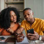 Zwei Personen sitzen gemeinsam an einem Tisch und schauen auf ein Smartphone. Eine Person h&auml;lt das Ger&auml;t, die andere zeigt auf den Bildschirm. Beide haben Notizen, einen Laptop und ein Getr&auml;nk vor sich.