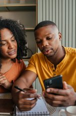 Reise-Apps, So planst du entspannt deinen Urlaub Zwei Personen sitzen gemeinsam an einem Tisch und schauen auf ein Smartphone. Eine Person hält das Gerät, die andere zeigt auf den Bildschirm. Beide haben Notizen, einen Laptop und ein Getränk vor sich.