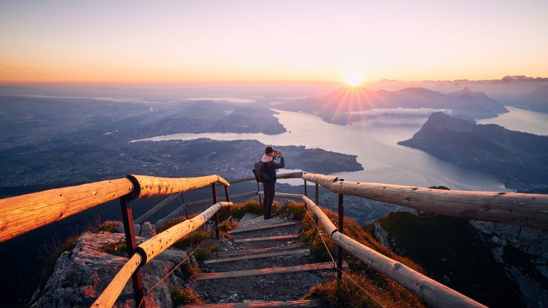 Schöne Urlaubsfotos machen, Die besten Tipps für tolle Fotos auf Reisen Eine Person steht vor einer beeindruckenden Seen- und Bergelandschaft bei Sonnenuntergang und hält ihre Kamera in den Händen.