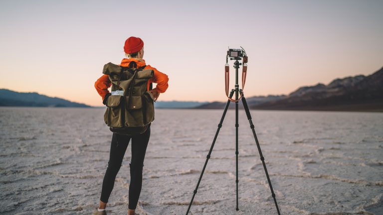 Eine Person steht in einer w&uuml;stenartigen Landschaft. Auf dem R&uuml;cken tr&auml;gt sie einen Fotorucksack, ihre Kamera steht auf einem Stativ.