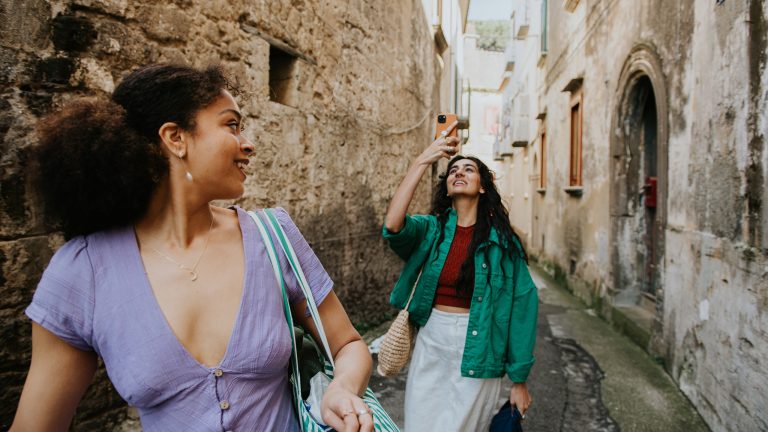 Zwei Personen befinden sich in einer engen steinernen Gasse. Eine Person nimmt ein Foto mit ihrem Smartphone auf, die andere blickt dabei &uuml;ber ihre Schulter.