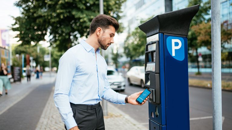 Eine Person steht an einem Parkscheinautomaten in einer Stadt und h&auml;lt ein Smartphone in der Hand. Im Hintergrund sind geparkte Autos, Gehweg und Geb&auml;ude zu sehen.