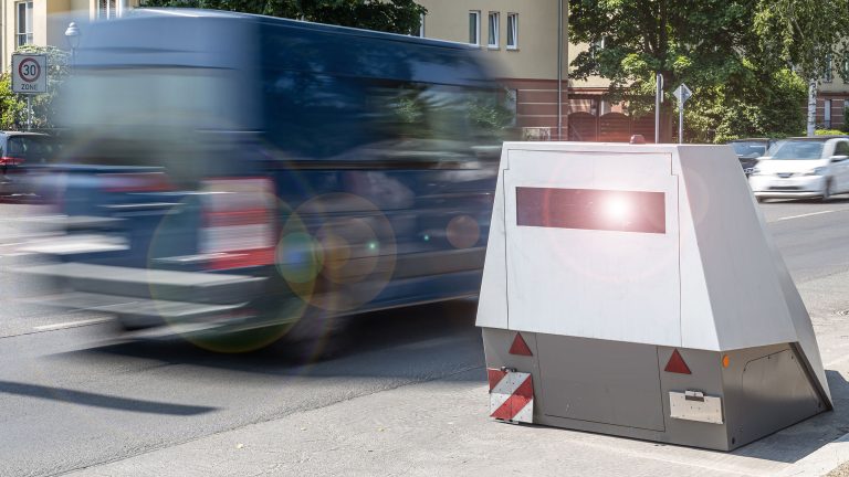 Ein station&auml;rer Blitzer steht am Stra&szlig;enrand, w&auml;hrend ein Fahrzeug mit hoher Geschwindigkeit vorbeif&auml;hrt. Im Hintergrund sind Geb&auml;ude und B&auml;ume zu sehen.