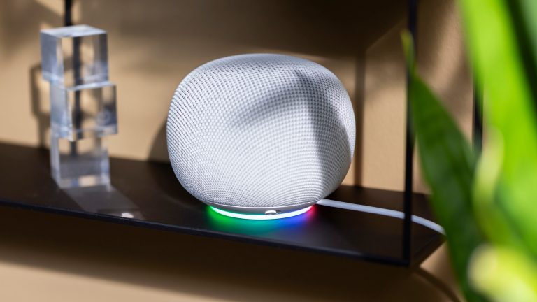 Google Home Speaker, Lautsprecher mit Gemini ausprobiert Ein Google Home Speaker mit leuchtendem Lichtring auf einem schwarzen Regal.