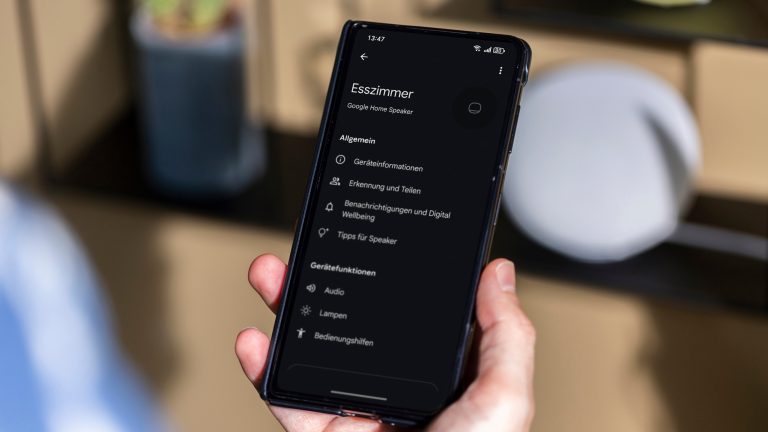 Google Home Speaker, Lautsprecher mit Gemini ausprobiert Eine Person hält ein Smartphone in der Hand, auf der die Google Home App geöffnet ist. Im Hintergrund steht ein Google Home Speaker.