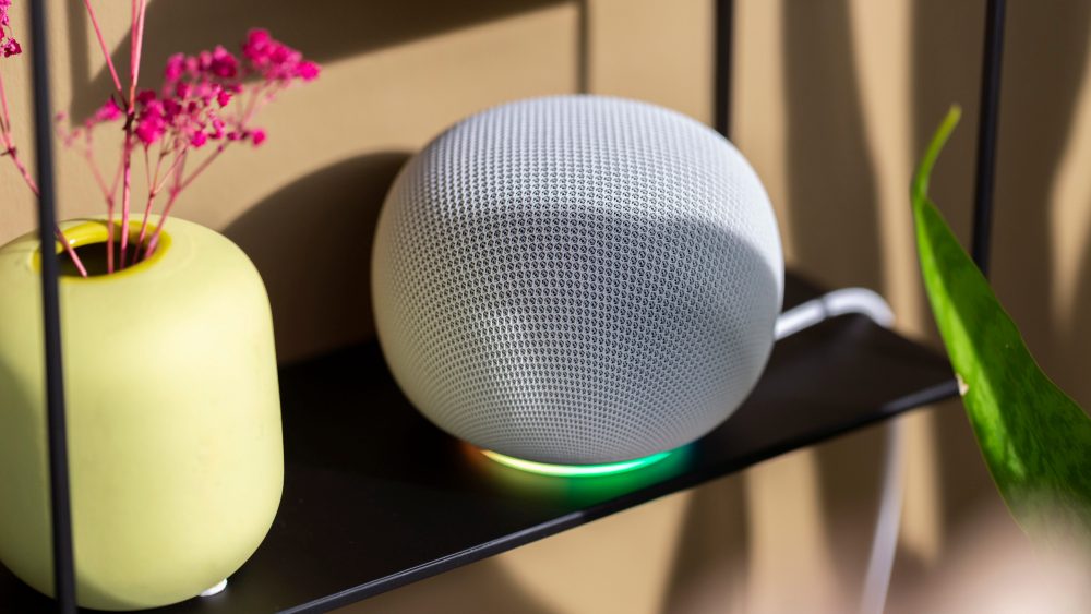 Ein Google Home Speaker in Wei&szlig; steht auf einem schwarzen Regal. Sein Lichtring leuchtet bunt.