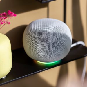 Ein Google Home Speaker in Wei&szlig; steht auf einem schwarzen Regal. Sein Lichtring leuchtet bunt.