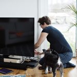 Eine Person kniet vor einem Fernseher und schlie&szlig;t Kabel an. Vor dem TV stehen eine Soundbar und weitere Ger&auml;te, daneben steht ein Hund.