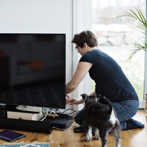 Eine Person kniet vor einem Fernseher und schlie&szlig;t Kabel an. Vor dem TV stehen eine Soundbar und weitere Ger&auml;te, daneben steht ein Hund.