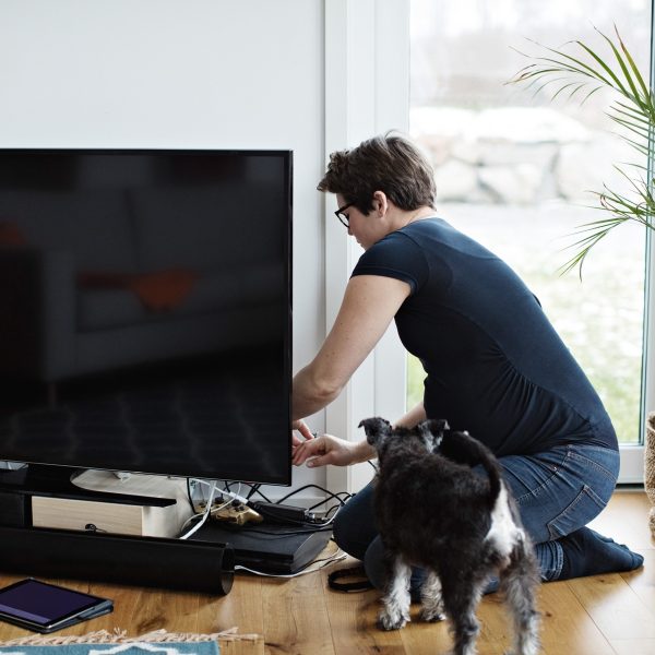 Eine Person kniet vor einem Fernseher und schlie&szlig;t Kabel an. Vor dem TV stehen eine Soundbar und weitere Ger&auml;te, daneben steht ein Hund.