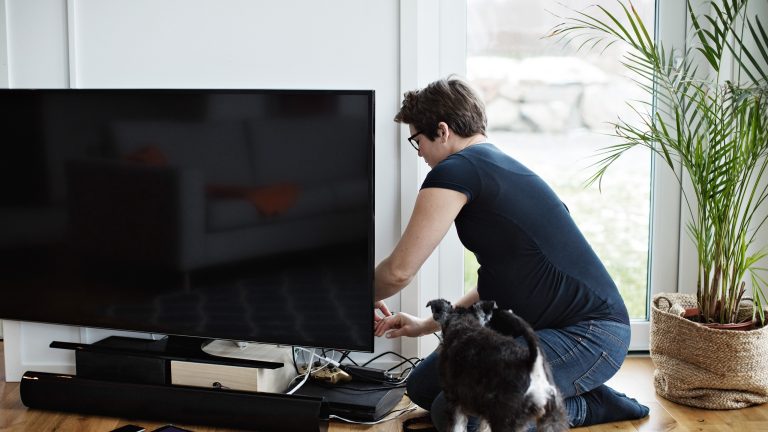 Eine Person kniet vor einem Fernseher und schlie&szlig;t Kabel an. Vor dem TV stehen eine Soundbar und weitere Ger&auml;te, daneben steht ein Hund.