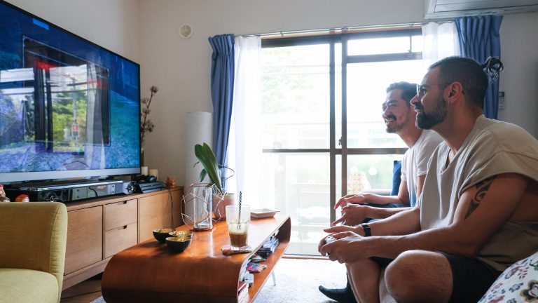 Zwei Personen sitzen vor einem Fernseher im Wohnzimmer und blicken auf den Bildschirm.