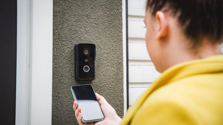 Outdoor-Kameras, So findest du das passende Modell Eine Person hält ein Smartphone vor eine Video-Türsprechanlage, die an einer Hauswand montiert ist.