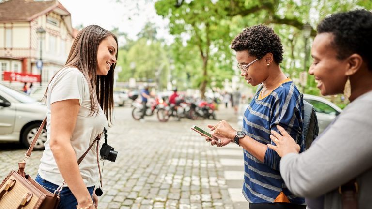 Urlaub clever planen, Die besten Apps zur Reise-Vorbereitung Drei Personen stehen zusammen auf einem Platz und schauen auf ein Smartphone in einer Hand. Im Hintergrund sind Gebäude und Bäume zu sehen.