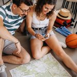 Zwei Personen sitzen auf dem Boden und schauen gemeinsam auf ein Smartphone. Vor ihnen liegt eine Karte, daneben stehen Reisetaschen und weitere Reiseutensilien.