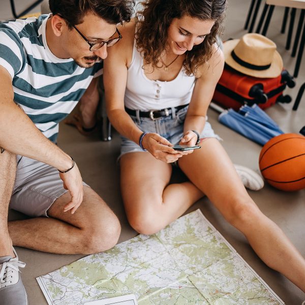 Zwei Personen sitzen auf dem Boden und schauen gemeinsam auf ein Smartphone. Vor ihnen liegt eine Karte, daneben stehen Reisetaschen und weitere Reiseutensilien.