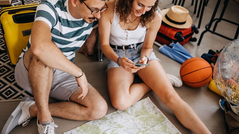 Zwei Personen sitzen auf dem Boden und schauen gemeinsam auf ein Smartphone. Vor ihnen liegt eine Karte, daneben stehen Reisetaschen und weitere Reiseutensilien.