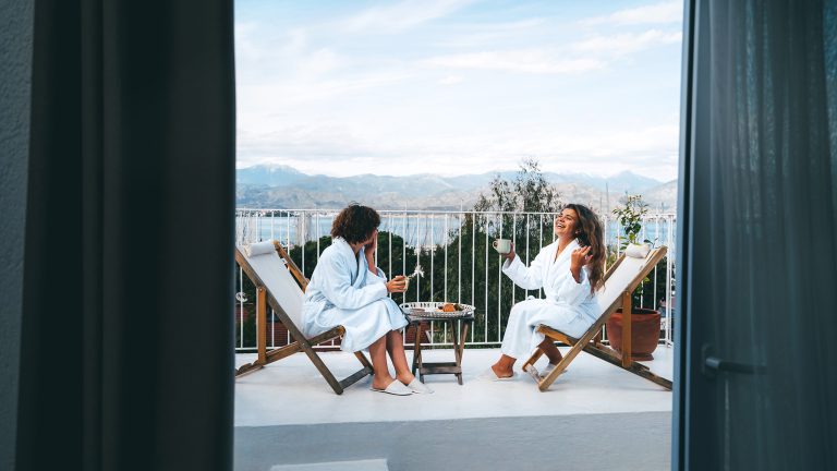 Zwei Personen sitzen in wei&szlig;en Badem&auml;nteln auf Liegest&uuml;hlen auf einer Terrasse mit Bergblick. Zwischen ihnen steht ein kleiner Tisch.