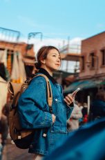 Die besten Apps für unterwegs, So wird dein Urlaub noch entspannter Eine Person mit Rucksack steht in einer belebten Gasse und hält ein Smartphone in der Hand. Im Hintergrund sind Marktstände, Gebäude und weitere Personen zu sehen.
