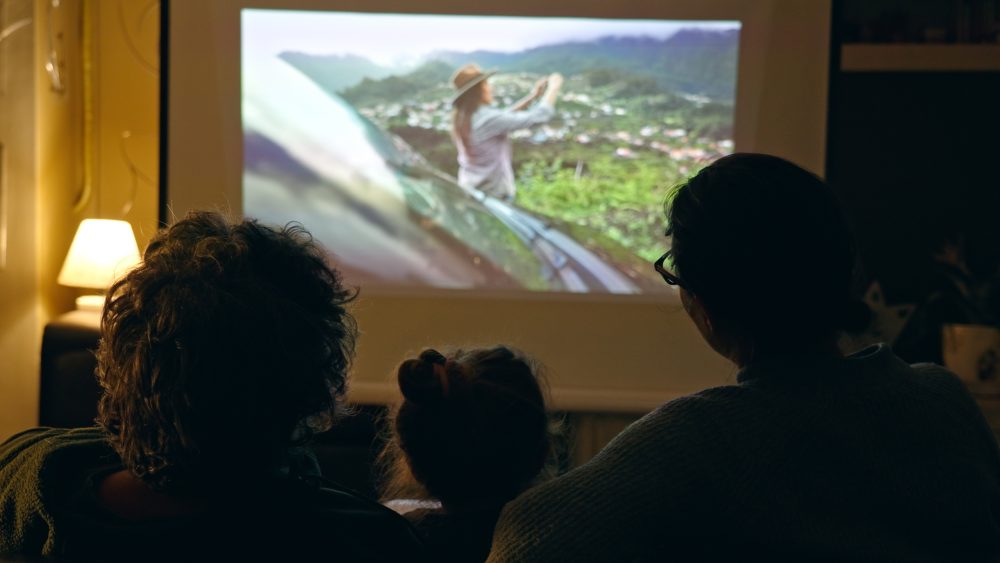 Mehrere Personen sitzen im Wohnzimmer vor einer Leinwand und sehen sich einen Film an. Der Raum ist abgedunkelt und die Personen sind von hinten zu sehen.