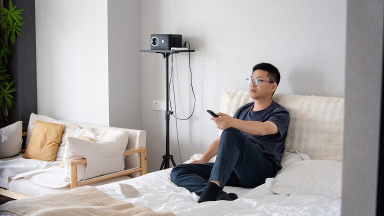 Eine Person sitzt auf einem Bett. In der linken Hand h&auml;lt sie eine Fernbedienung.
