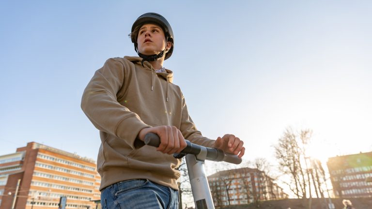 Person mit Helm auf E-Scooter bei Tageslicht in urbaner Umgebung; H&auml;nde am Lenker, Geb&auml;ude im Hintergrund.