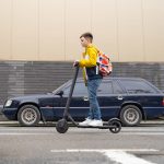 Jugendliche Person f&auml;hrt mit E-Scooter bei Tageslicht auf einer Stra&szlig;e zwischen Autos.