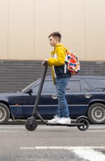 E‑Scooter für Kinder und Jugendliche, Was ist erlaubt? Jugendliche Person fährt mit E-Scooter bei Tageslicht auf einer Straße zwischen Autos.