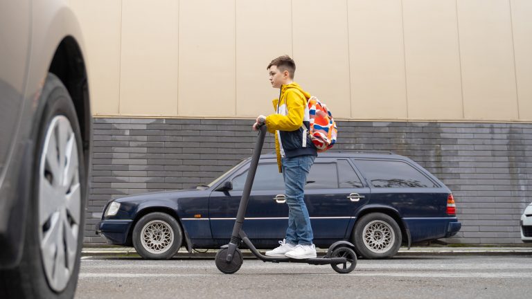 Jugendliche Person f&auml;hrt mit E-Scooter bei Tageslicht auf einer Stra&szlig;e zwischen Autos.