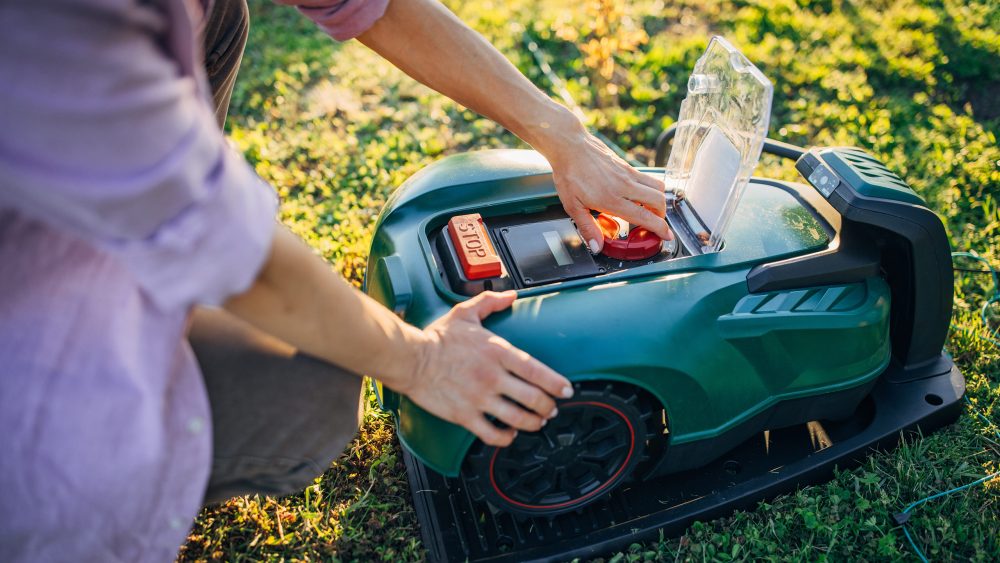 Eine Person kniet im Gras vor einem M&auml;hroboter und &ouml;ffnet diesen mit der linken Hand, um ihn zu reinigen und zu entleeren.