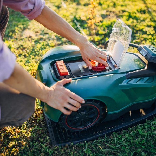 Eine Person kniet im Gras vor einem M&auml;hroboter und &ouml;ffnet diesen mit der linken Hand, um ihn zu reinigen und zu entleeren.