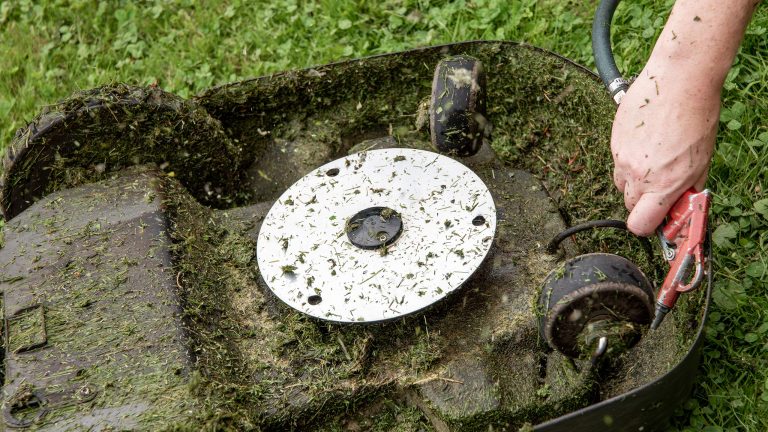 Ein M&auml;hroboter liegt kopf&uuml;ber im Gras und ist an der Unterseite ge&ouml;ffnet. Es ist die Hand einer Person zu sehen, die den M&auml;hroboter reinigt.