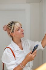 Eine Person stellt eine &Uuml;berwachungskamera auf eine Treppenstufe in einem Haus. Dabei h&auml;lt sie ein Smartphone in der Hand.