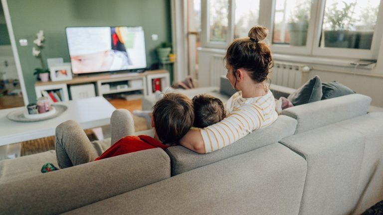 Drei Personen sitzen auf der Couch vor einem laufenden Fernseher. Sie wurden von hinten fotografiert.