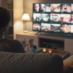 Eine Person sitzt auf einer Couch in einem abgedunkelten Wohnzimmer vor einem laufenden Fernseher. Es ist der R&uuml;cken der Person zu sehen.