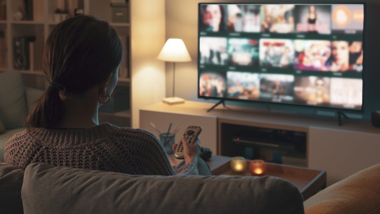 Eine Person sitzt auf einer Couch in einem abgedunkelten Wohnzimmer vor einem laufenden Fernseher. Es ist der R&uuml;cken der Person zu sehen.